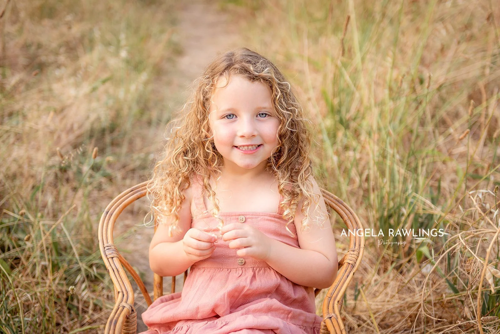 A young girl with curly blonde hair and blue eyes, wearing a pink dress, sitting on a rattan chair outdoors in a field of tall grass, smiling at the camera.