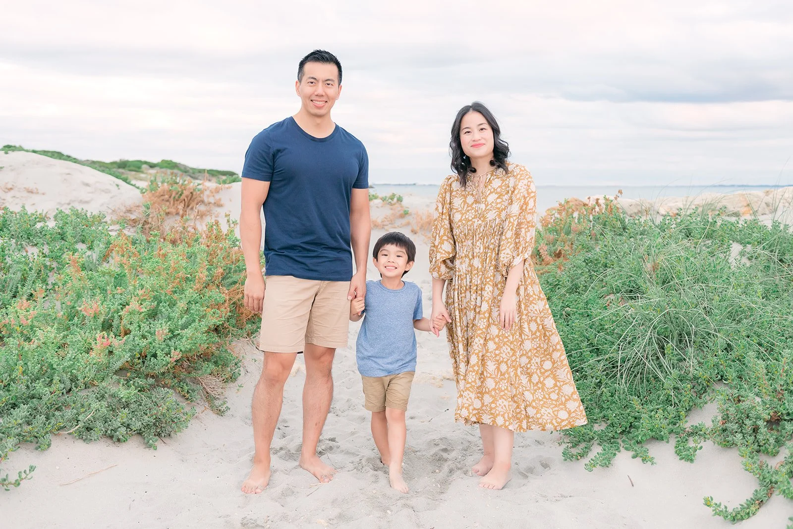 A family of three standing on a sandy beach with green bushes, a cloudy sky, and distant water in the background. The father and mother are holding hands with their young son.