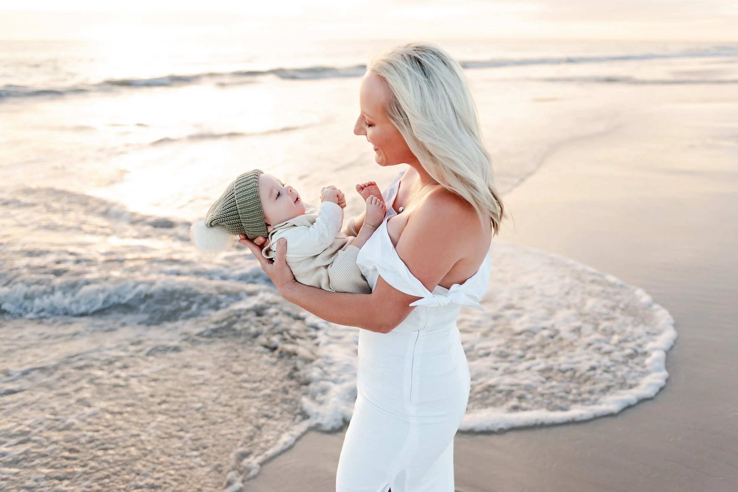 A woman with blonde hair holding a baby in a beige outfit and green knit hat at the beach, with the ocean and sunset in the background.