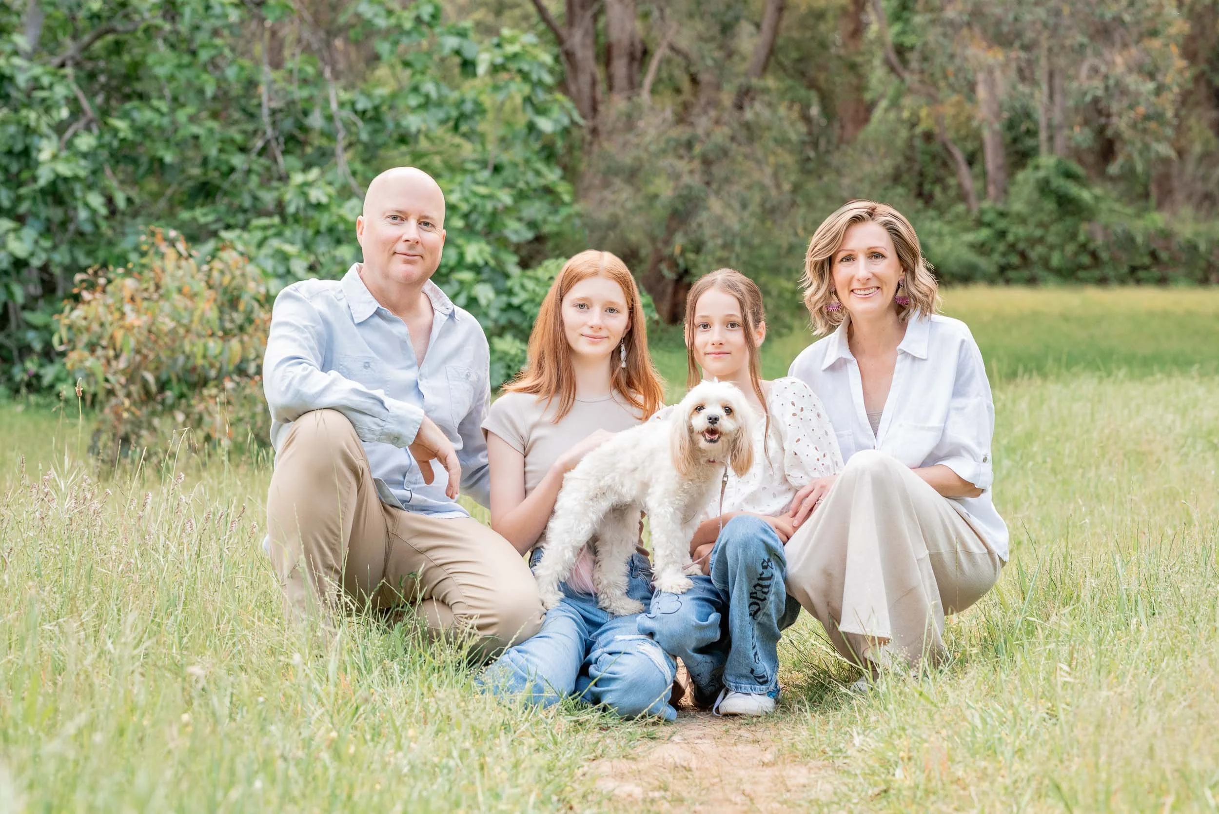 A family of four, including a man, a woman, two young girls, and a small dog, sitting on grass in a park with trees in the background.