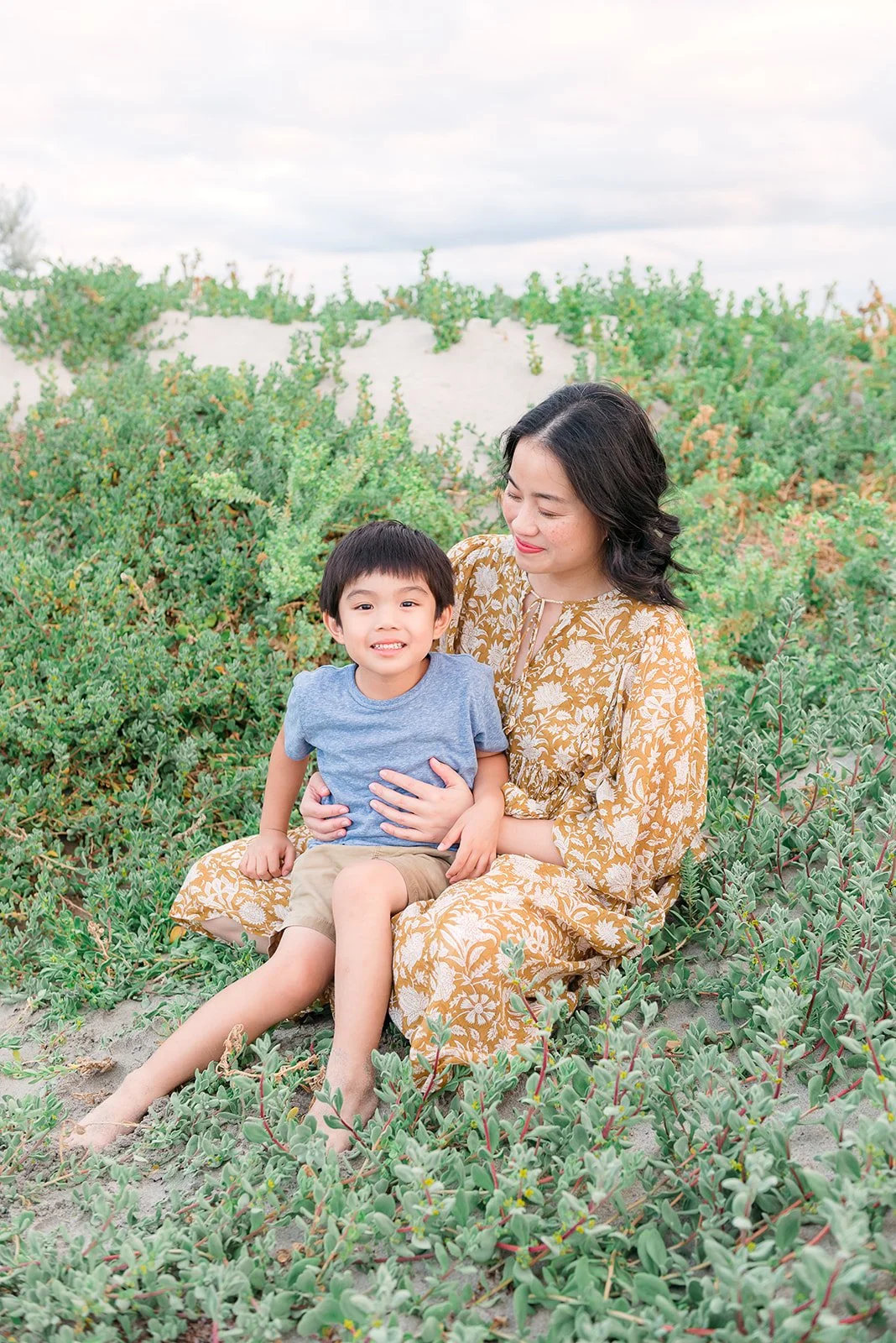 A woman and a young boy sitting on sandy ground surrounded by green plants with a cloudy sky overhead
