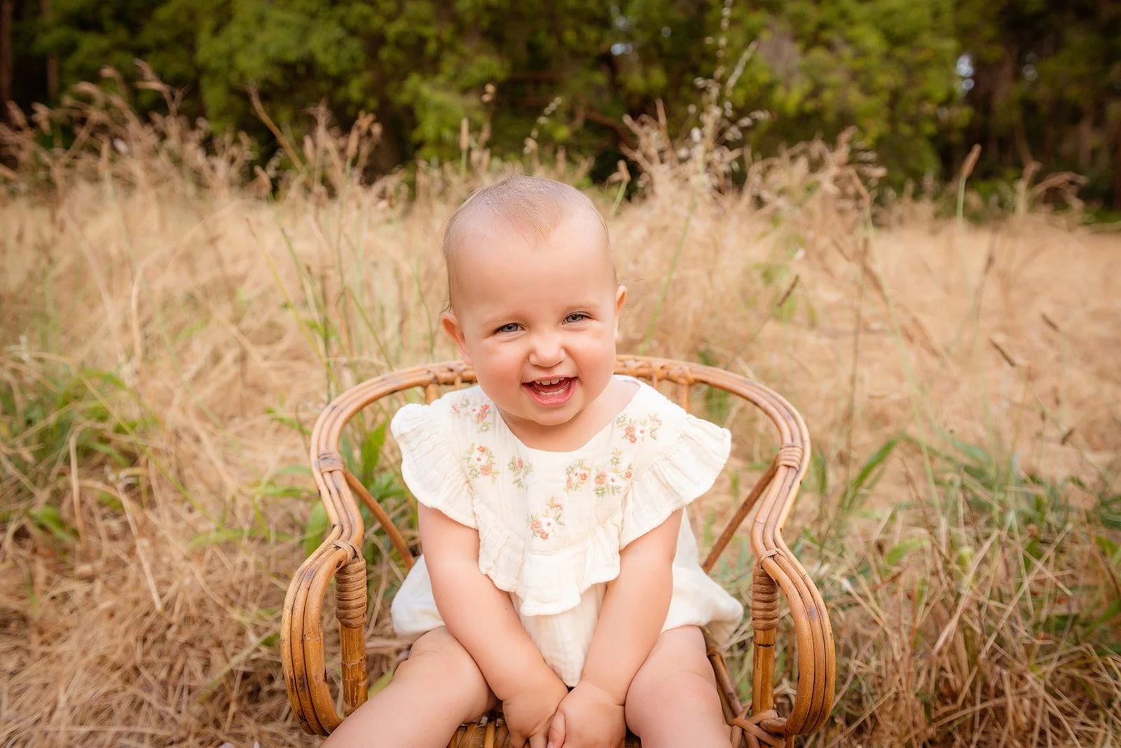 A smiling baby girl with blond hair, sitting in a wicker chair outdoors in a grassy field with tall, dry grass and trees in the background.