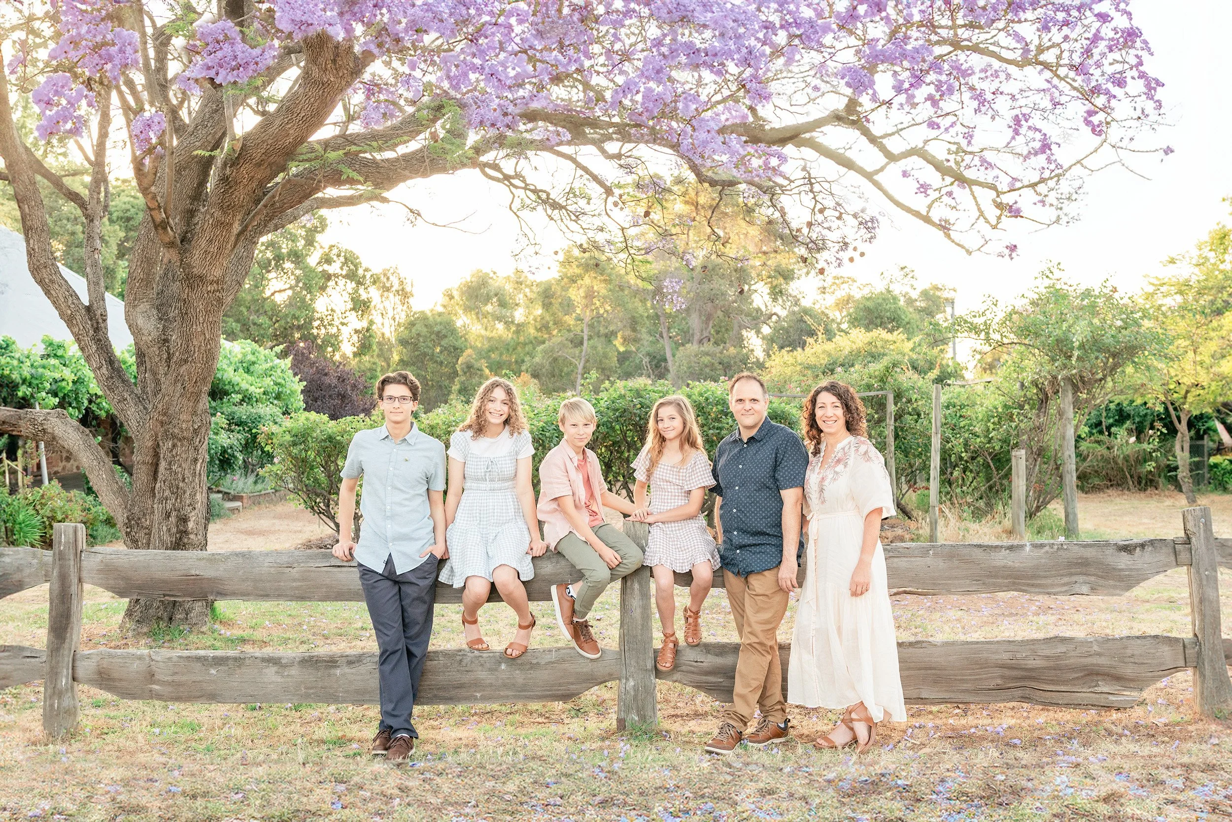 Family of six standing and sitting outdoors under a large purple-flowered tree during sunset, with green trees and a wooden fence in the background.
