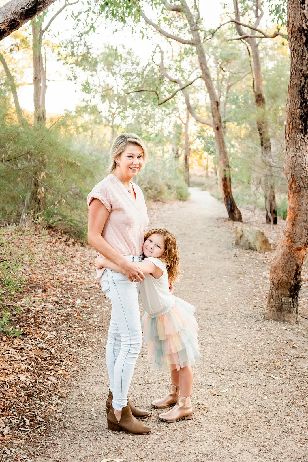 A woman and a young girl hugging on a dirt trail surrounded by trees in a forested area during sunset.