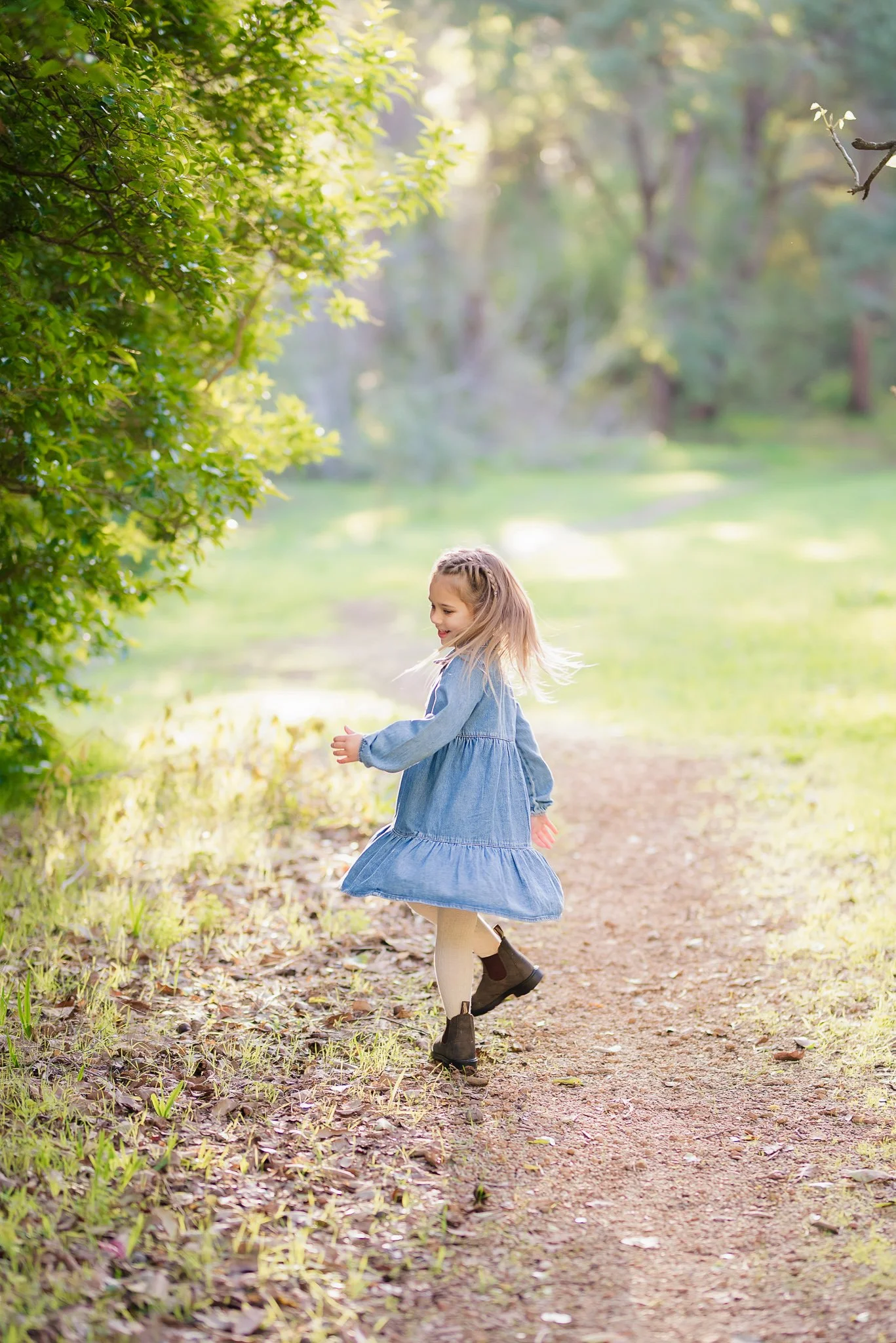 A young girl with long blonde hair, wearing a blue dress, beige tights, and dark boots, running along a dirt path in a green outdoor park or forest area during sunny weather.