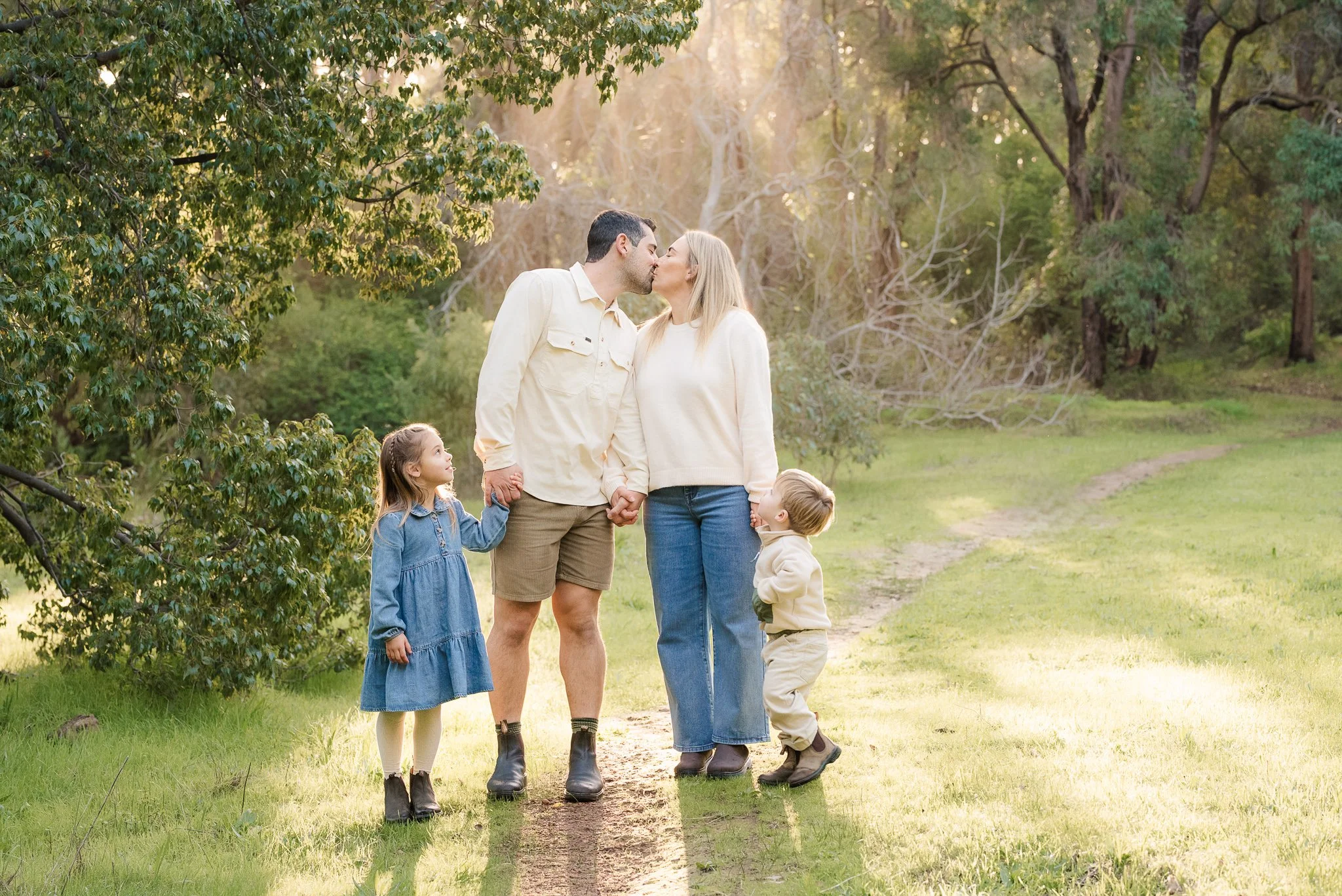 A family of four holding hands and kissing outdoors on a grassy trail with trees in the background.