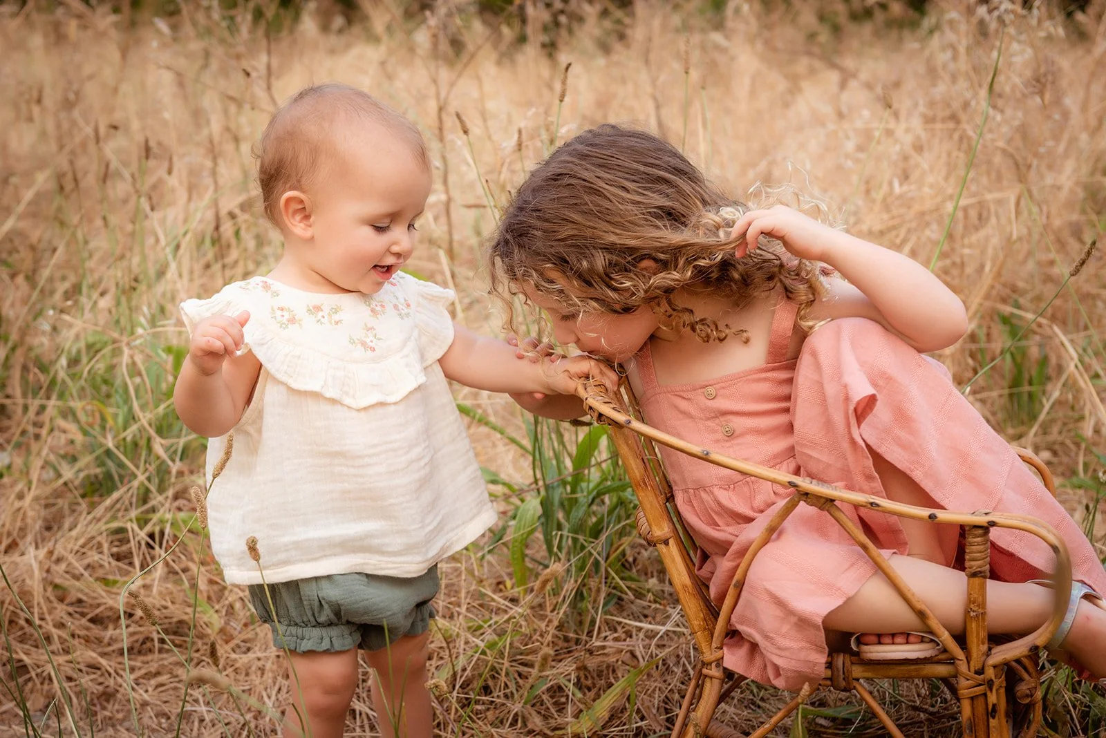 Two young girls playing outdoors in a grassy field, one standing and the other sitting in a wicker chair, engaging with each other.