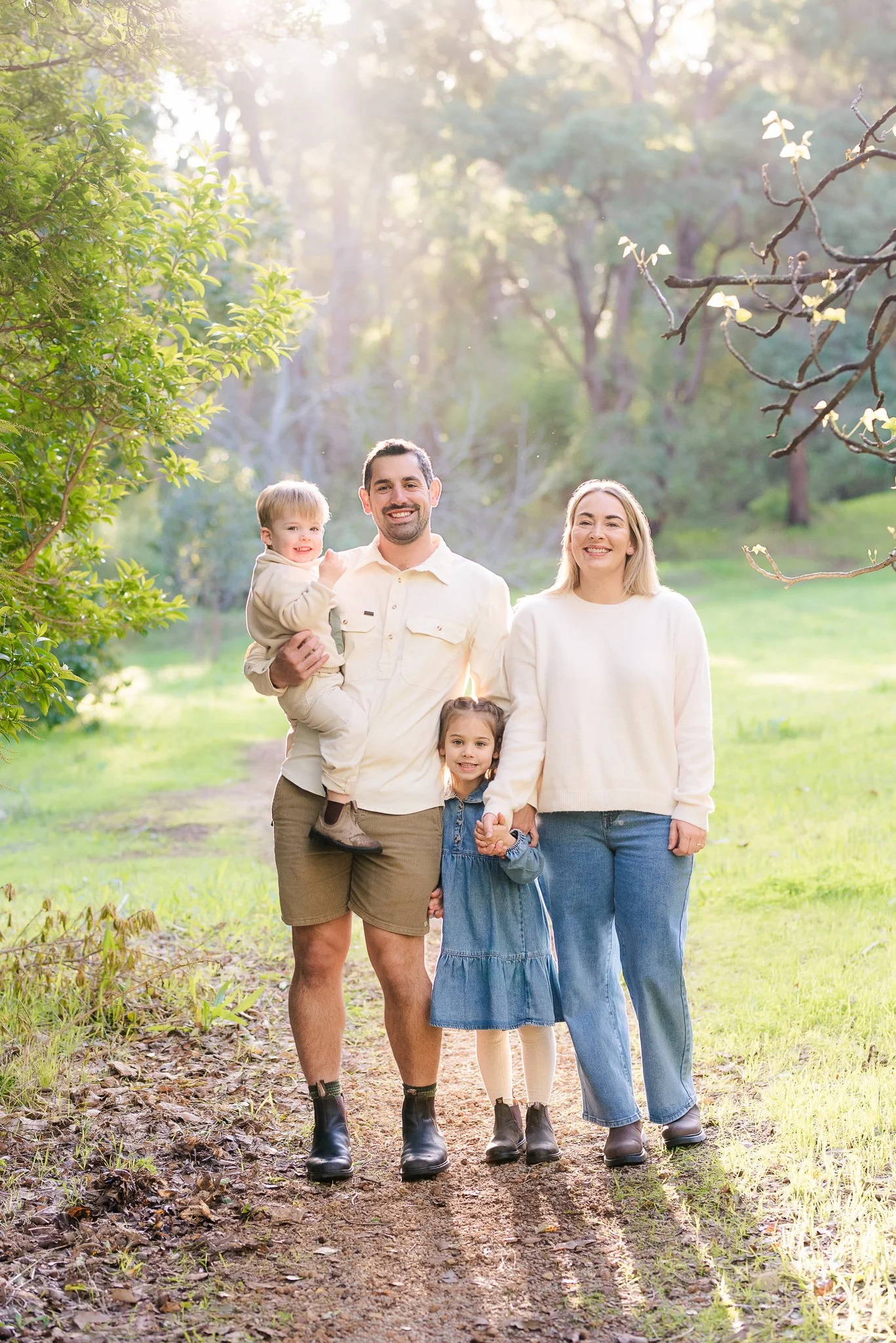 A family of five enjoying a walk outdoors in a wooded area, holding hands and smiling at the camera with sunlight filtering through the trees.