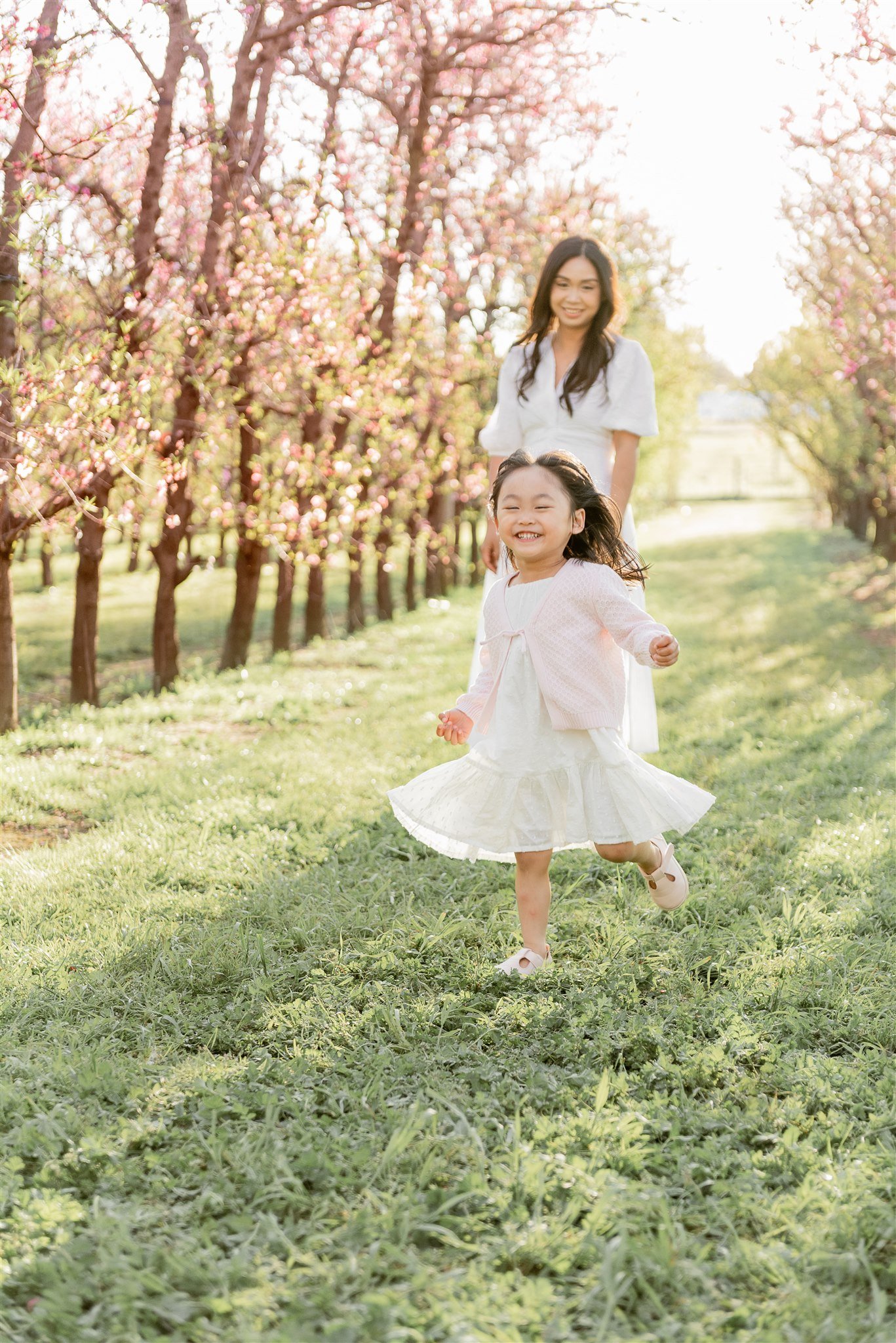 A young girl in a white dress and pink cardigan running through a grassy area with blooming pink trees, smiling joyfully, with a woman in a white dress following behind.