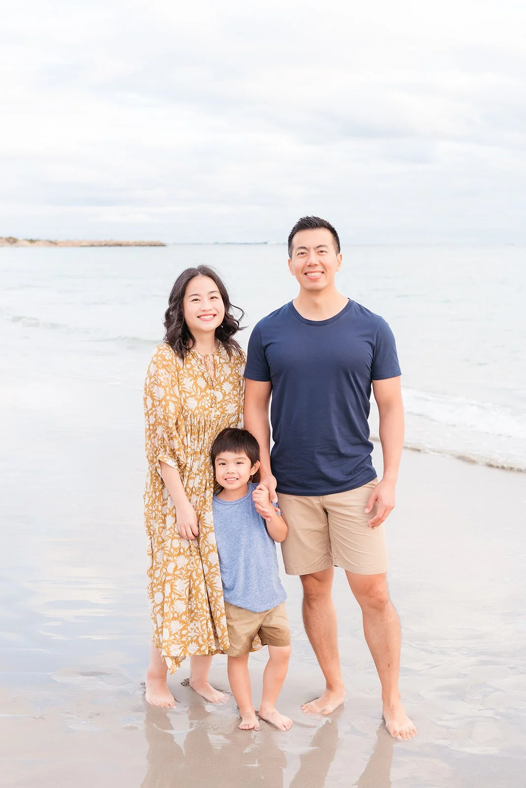 A family of three standing on a beach with the ocean in the background. The woman has shoulder-length dark hair and is wearing a yellow floral dress. The man has short dark hair and is dressed in a navy t-shirt and khaki shorts. Their young son is st