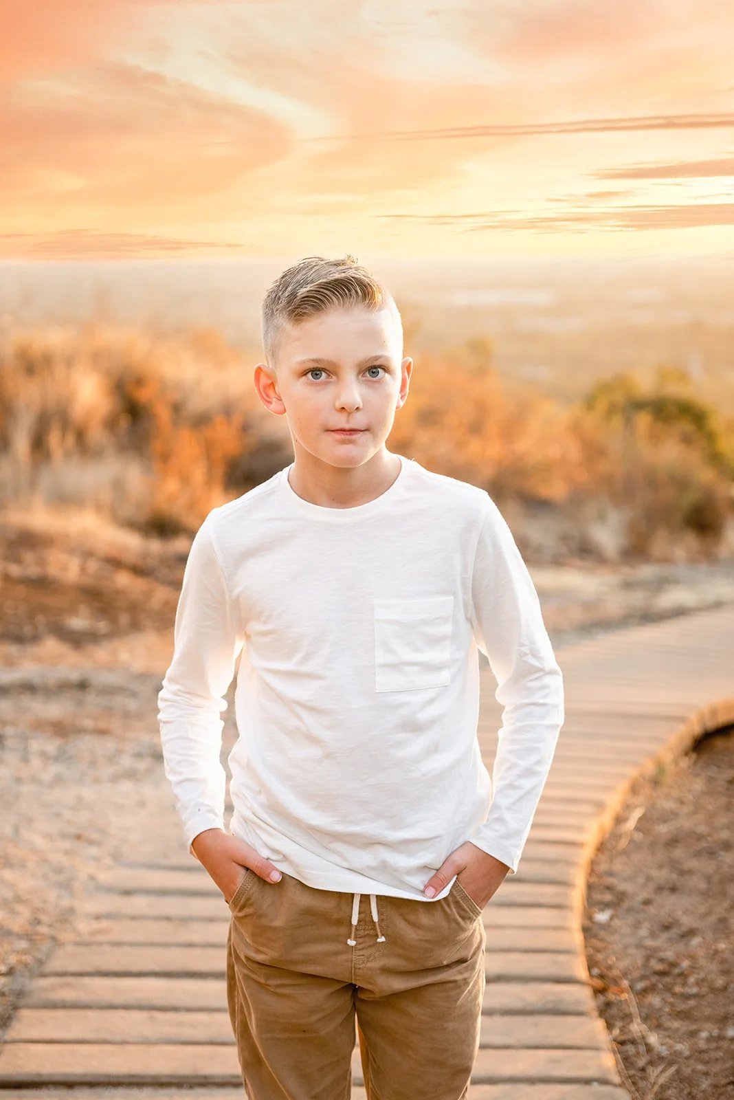 A young boy with short blonde hair and blue eyes wearing a white long-sleeve shirt and tan pants standing on a winding wooden trail during sunset, with orange and yellow sky in the background.