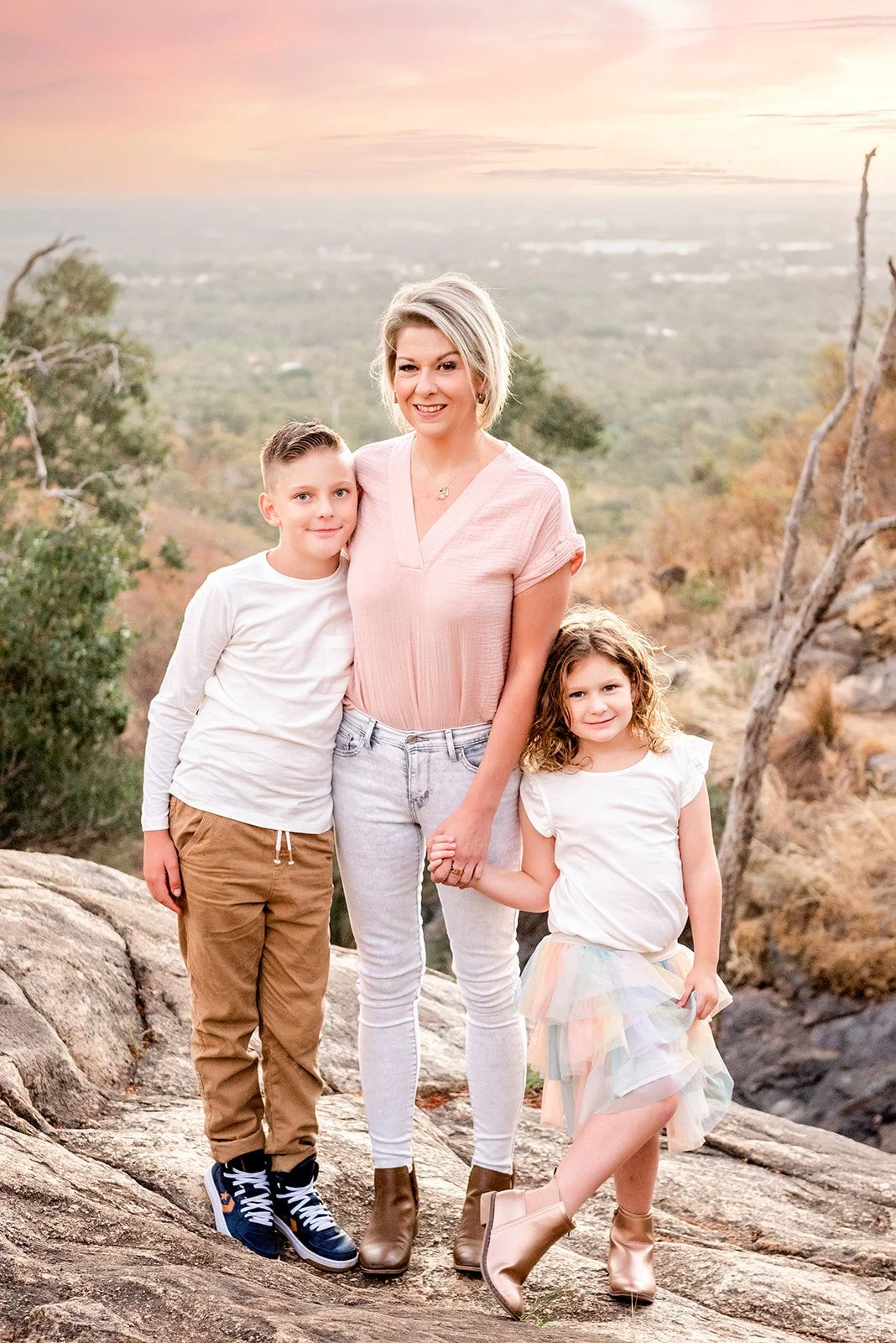 A woman with short blonde hair and two children standing outdoors on a rocky hillside at sunset. The woman is wearing a pink top and white jeans, holding the hand of a young girl with curly hair in a pastel tutu skirt. A young boy in a white shirt an