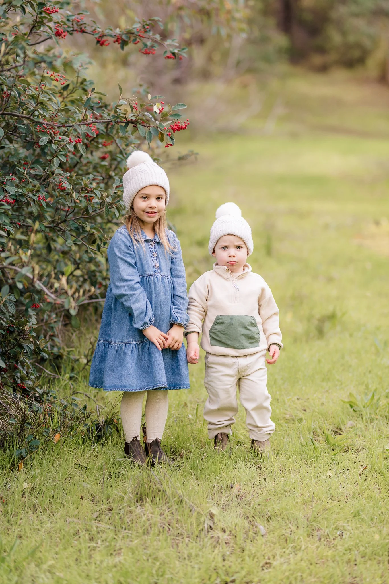 Two children standing on grass outdoors near a bush with red berries. The girl on the left wears a denim dress, beige tights, brown boots, and a white knit hat with a pompom. The boy on the right wears a cream hoodie with a green pocket, beige pants,