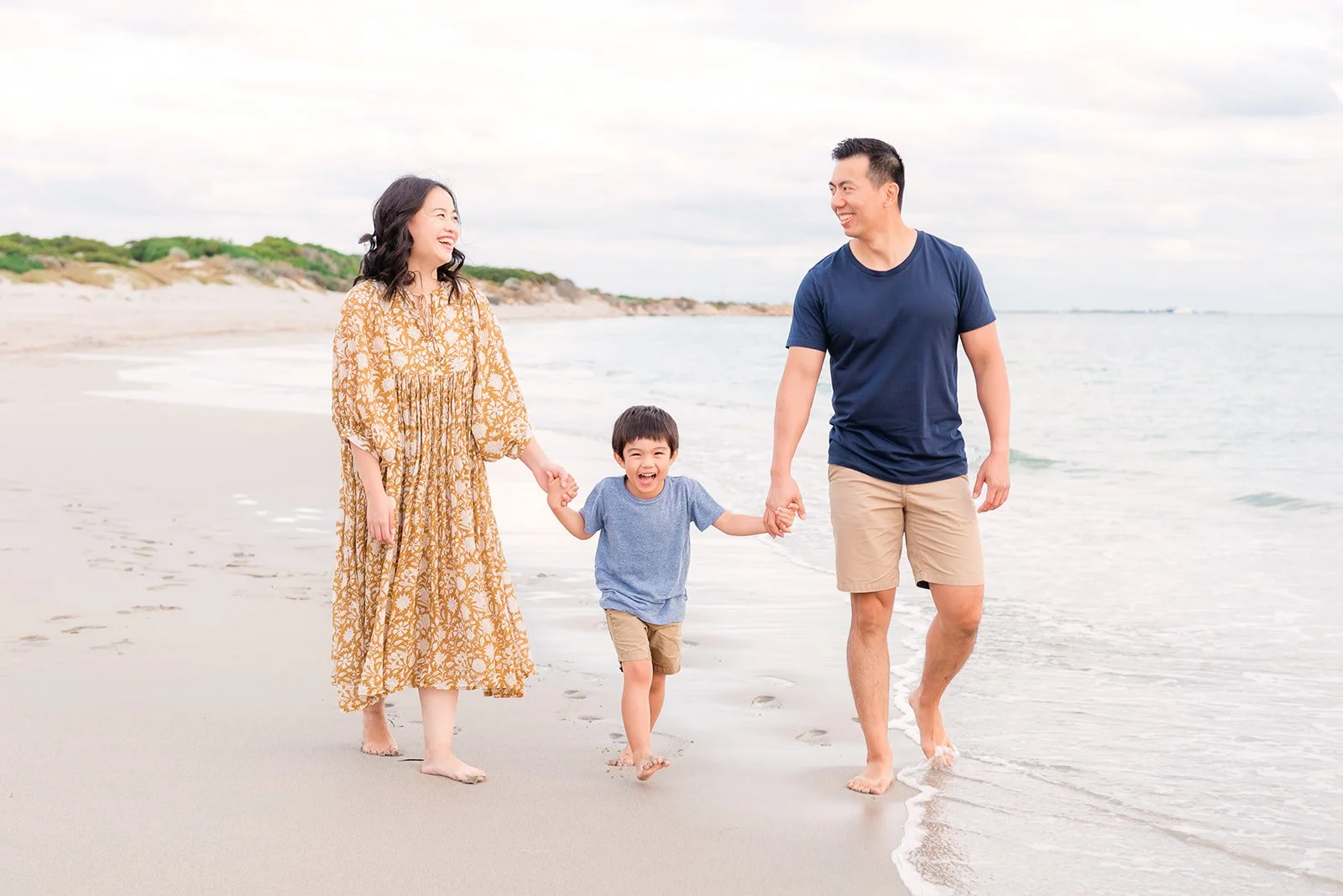 A happy family of three walking on the beach, holding hands, with the ocean in the background.