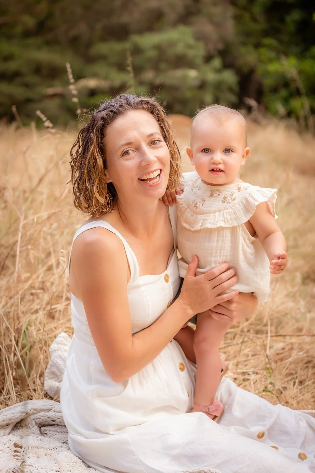 A woman with curly hair in a white dress holding a young girl with blonde hair in a white dress, sitting in a field of tall, dry grass with trees in the background.