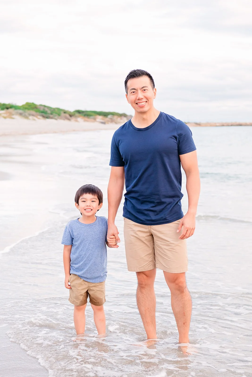A man and a boy holding hands while standing in the water at the beach, smiling at the camera.