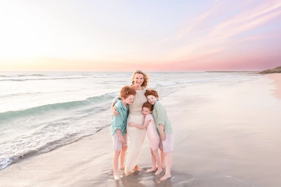 A woman and three children hugging on a beach at sunset.