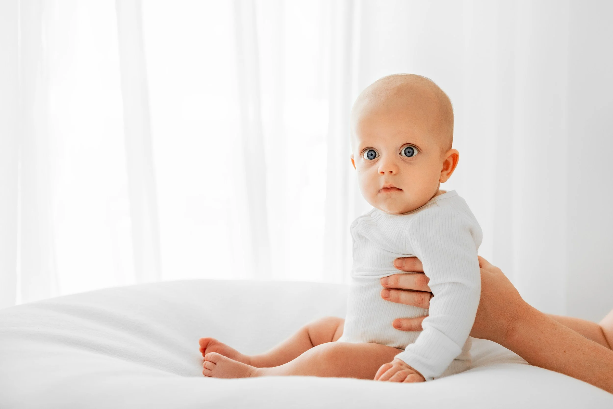 An infant with big blue eyes and a white long-sleeved outfit, sitting on a white surface in front of a softly lit background.