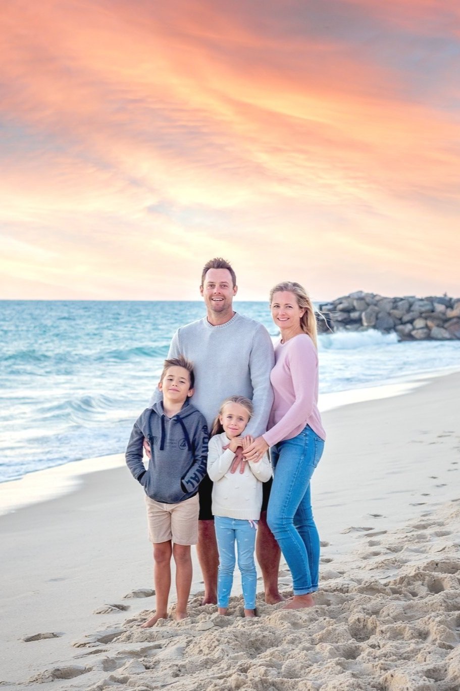 A family of four on a beach at sunset, standing on sand near the ocean with a rocky jetty in the background. The family includes a man, woman, and two children, all smiling and facing the camera.