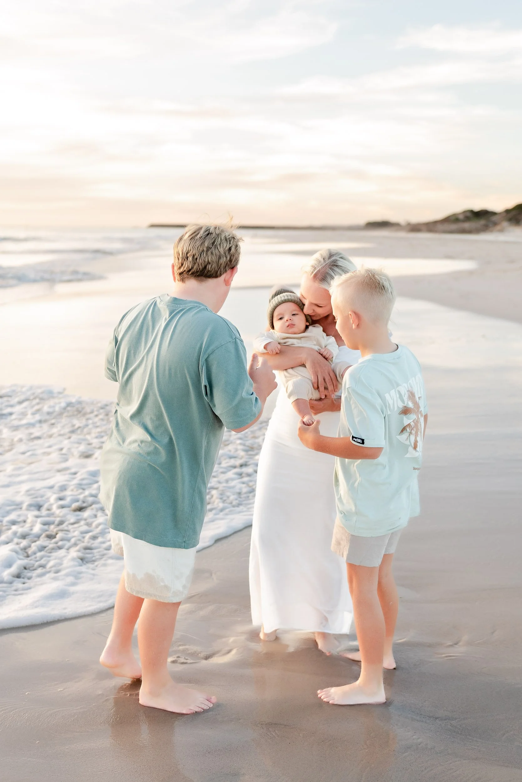 A family on the beach during sunset, with a woman holding a baby and two boys standing near her, all barefoot and engaged in a moment of affection.