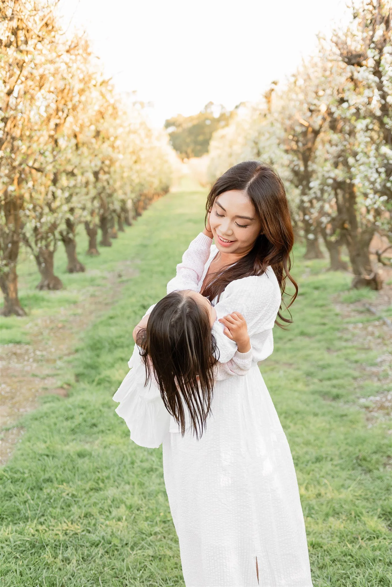 A woman wearing a white dress holding a young girl in her arms, mother is smiling at her daughter, in a lush green orchard with rows of white cherry blossoms in the background, during golden hour.