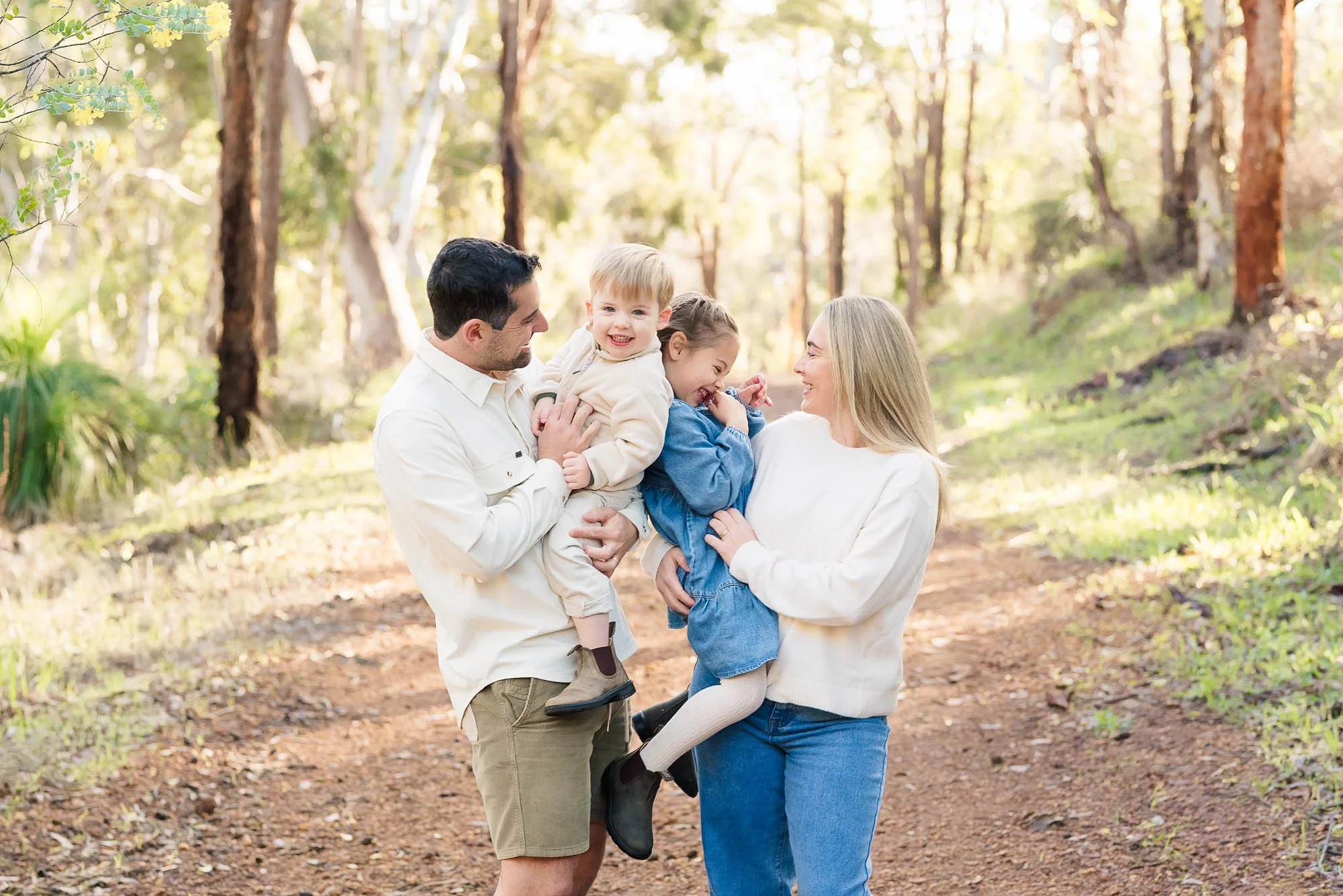 A happy family of four enjoying time outdoors in a wooded area during daytime, with the father and mother holding their two young children and all smiling.