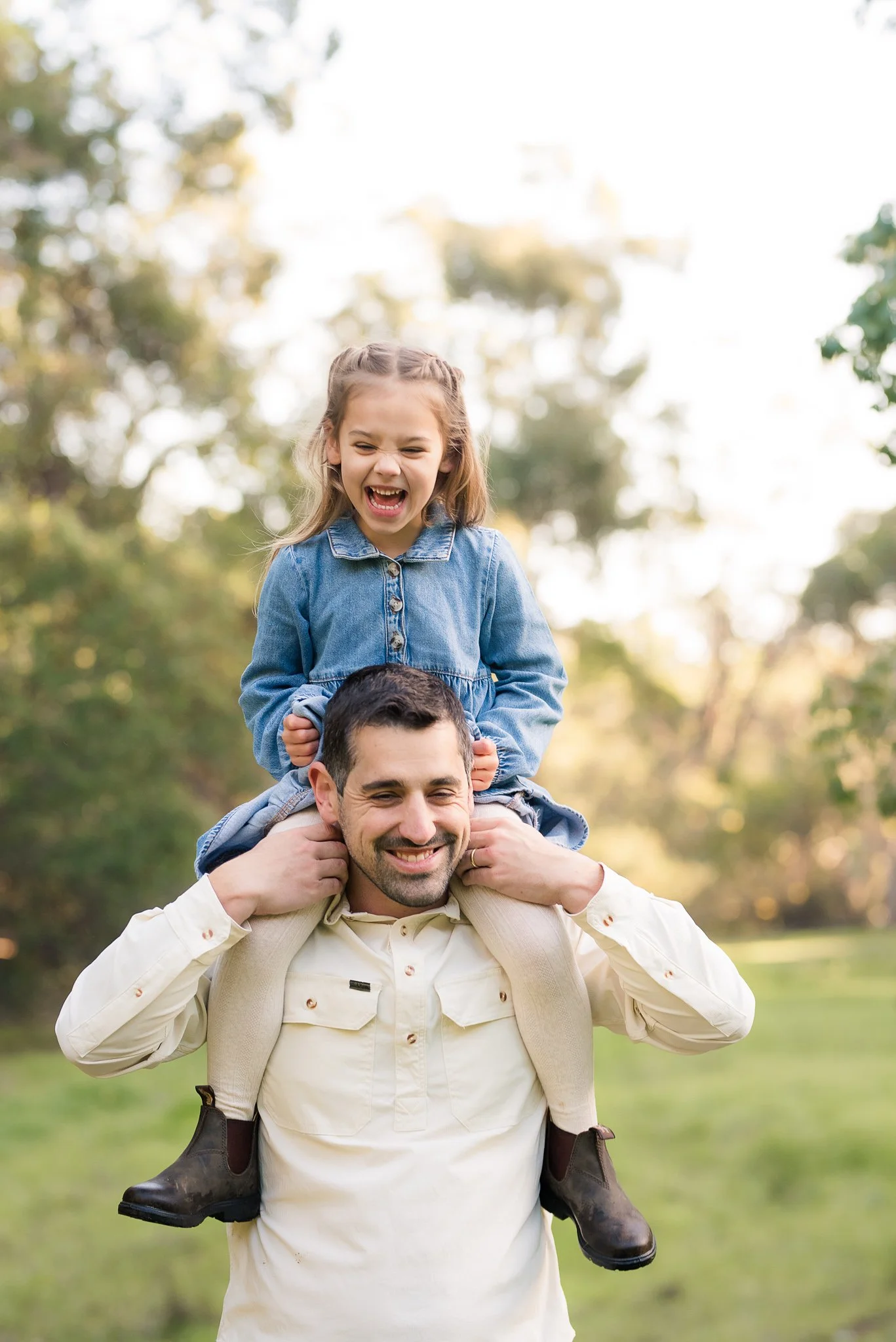 A man carrying a young girl on his shoulders outside in a park with trees, both smiling and enjoying the moment.