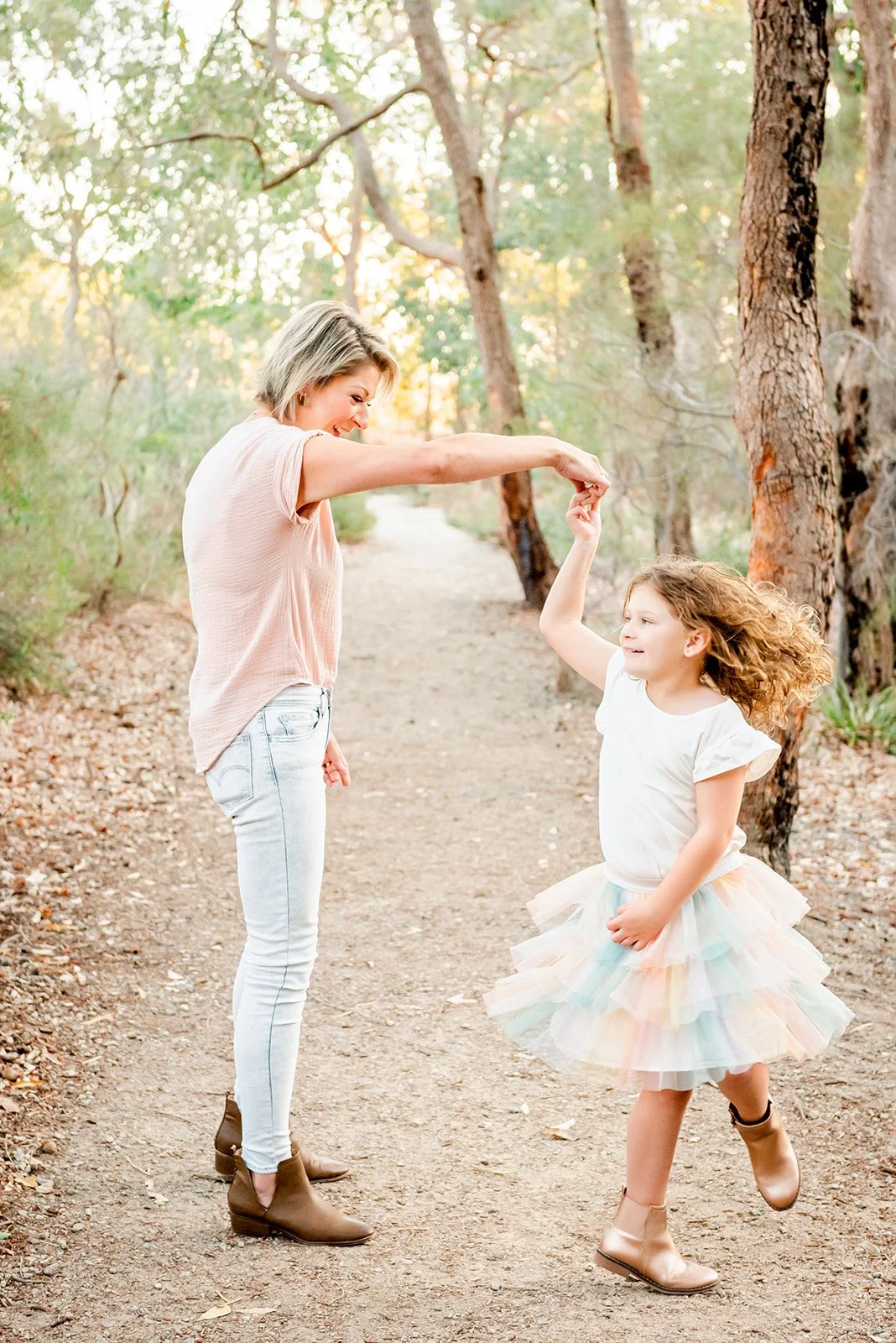 A woman and a young girl are dancing and twirling on a wooded dirt path, holding hands and smiling, with trees and sunlight in the background.