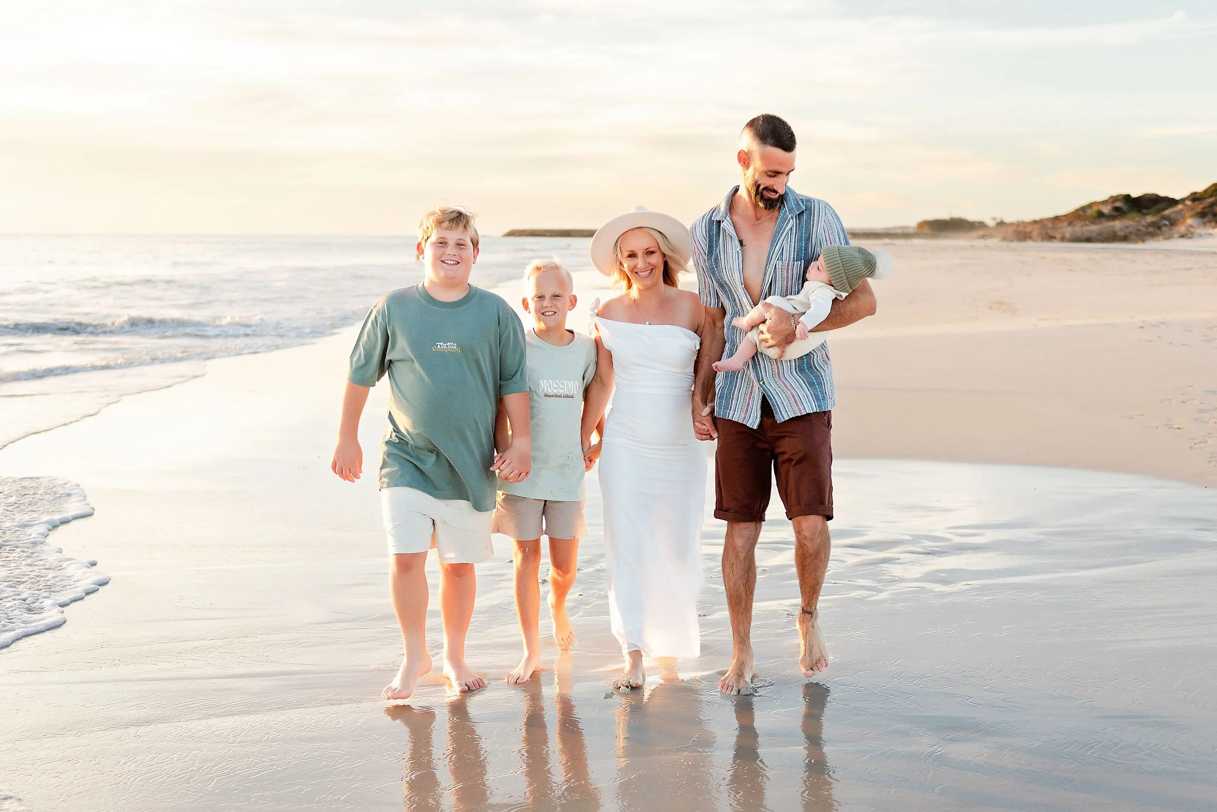 A happy family walking on the beach during sunset, holding hands, with the father holding a baby, and the mother wearing a white dress and hat.