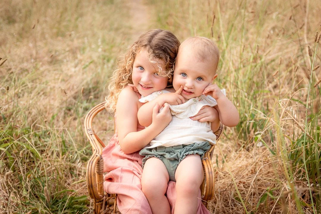 Two young children, a girl with curly hair and a baby, sitting together on a wicker chair in a field of tall grass.