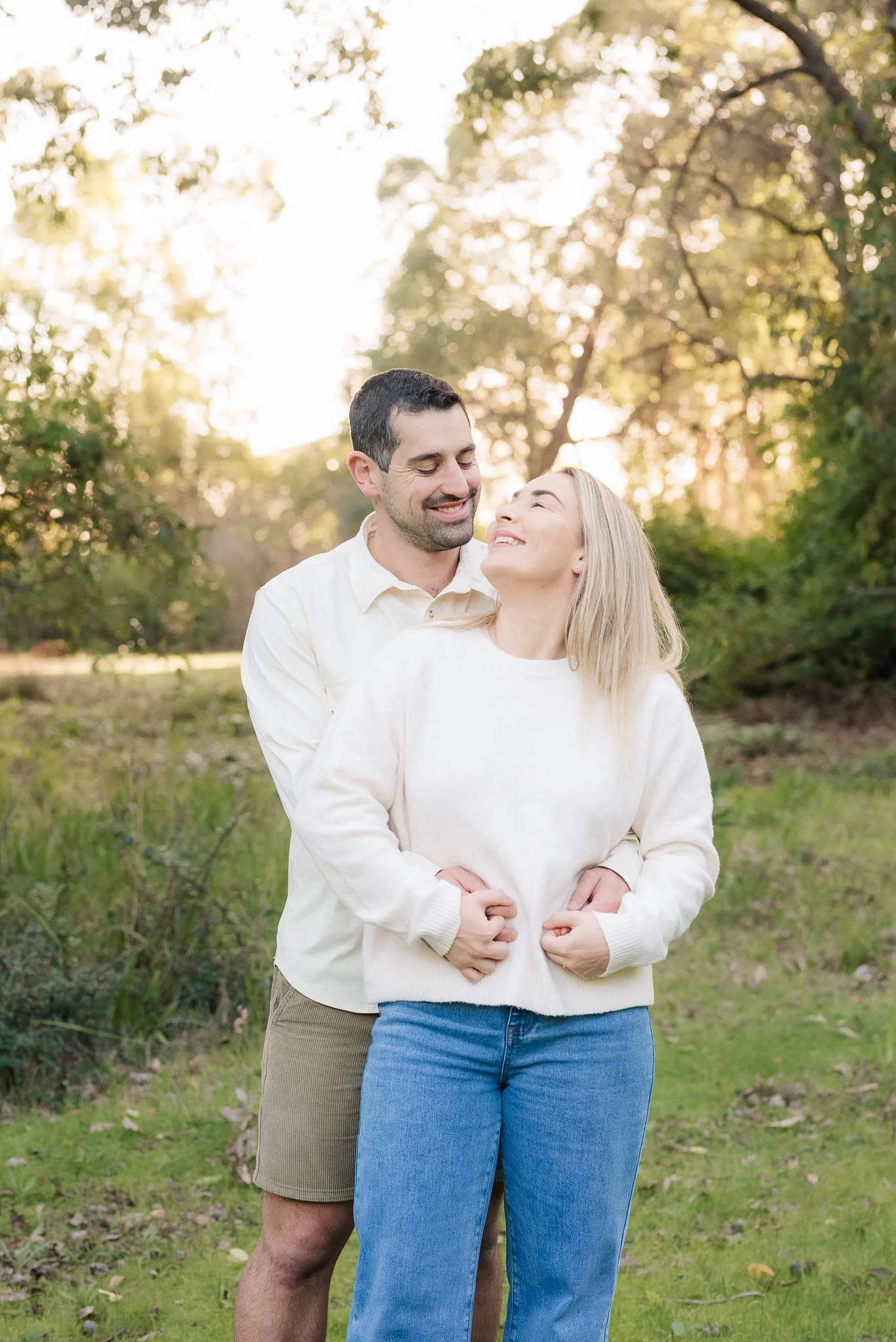 A couple standing outdoors in the Perth Hills, smiling and embracing each other, with sunlight filtering through the trees in the background.