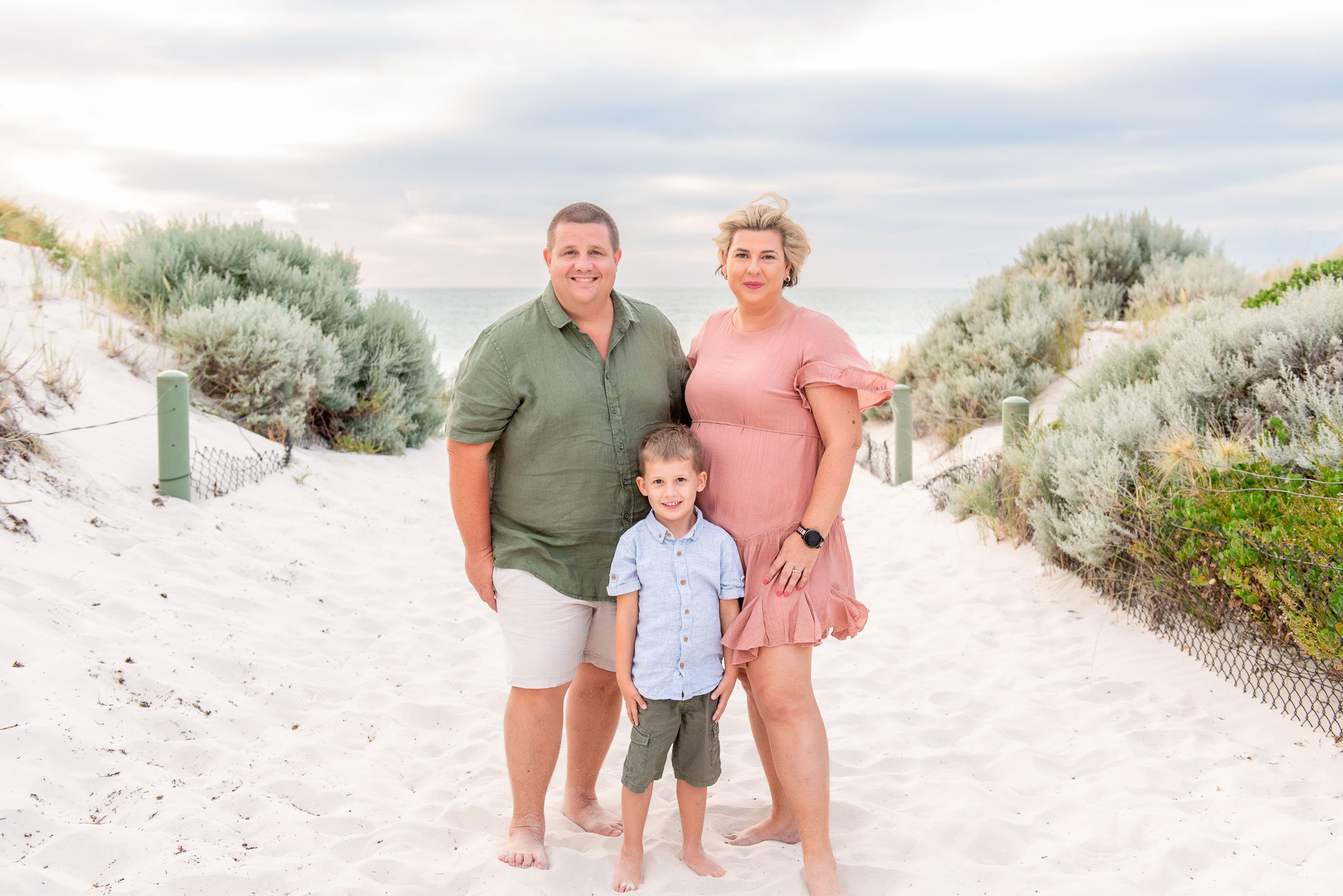 A family of three standing on a sandy beach with dune vegetation, ocean, and cloudy sky in background.