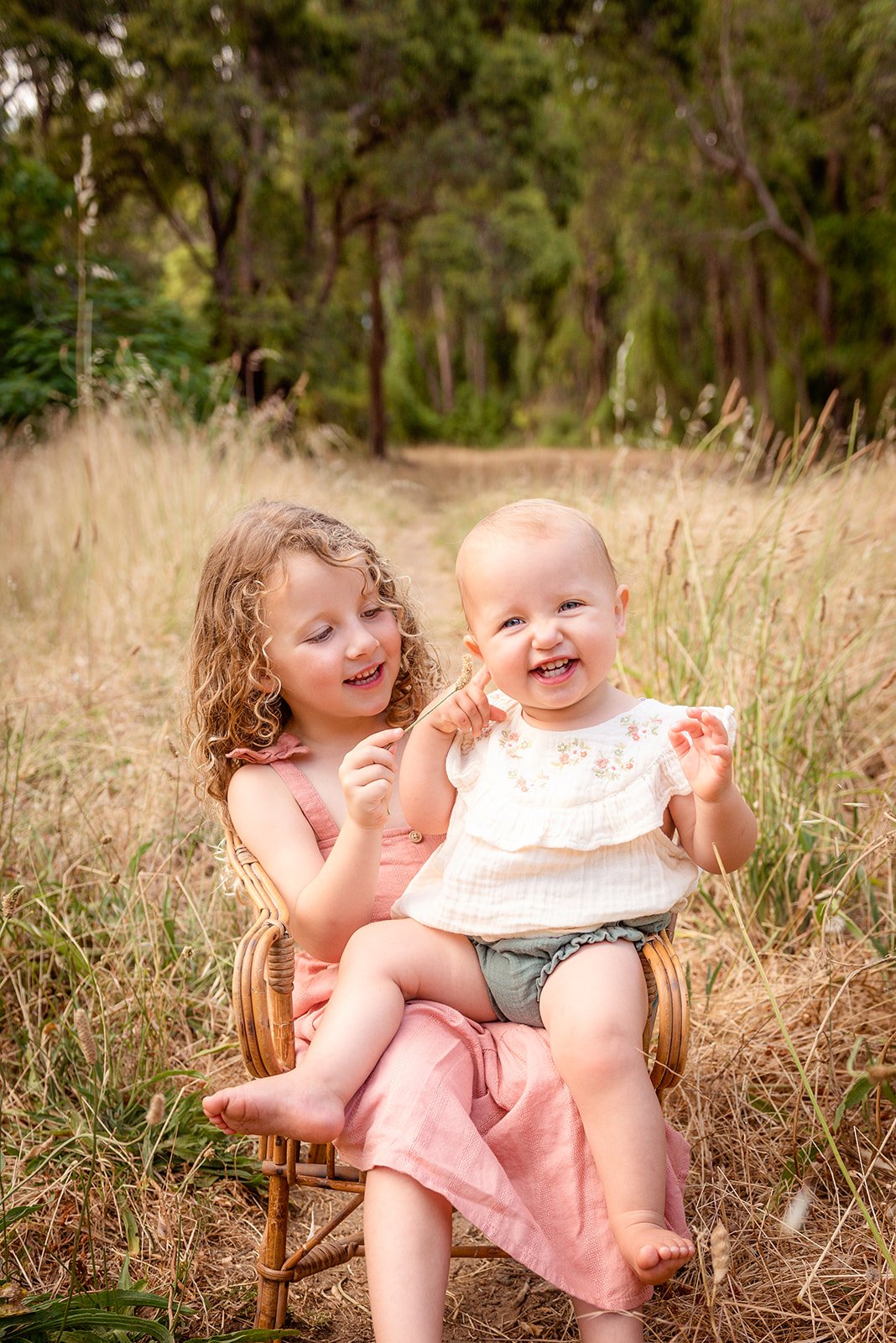 Two young girls, one with curly hair and the other with short, light-colored hair, sitting together on a wicker chair outdoors in a grassy, wooded area, smiling and playing.