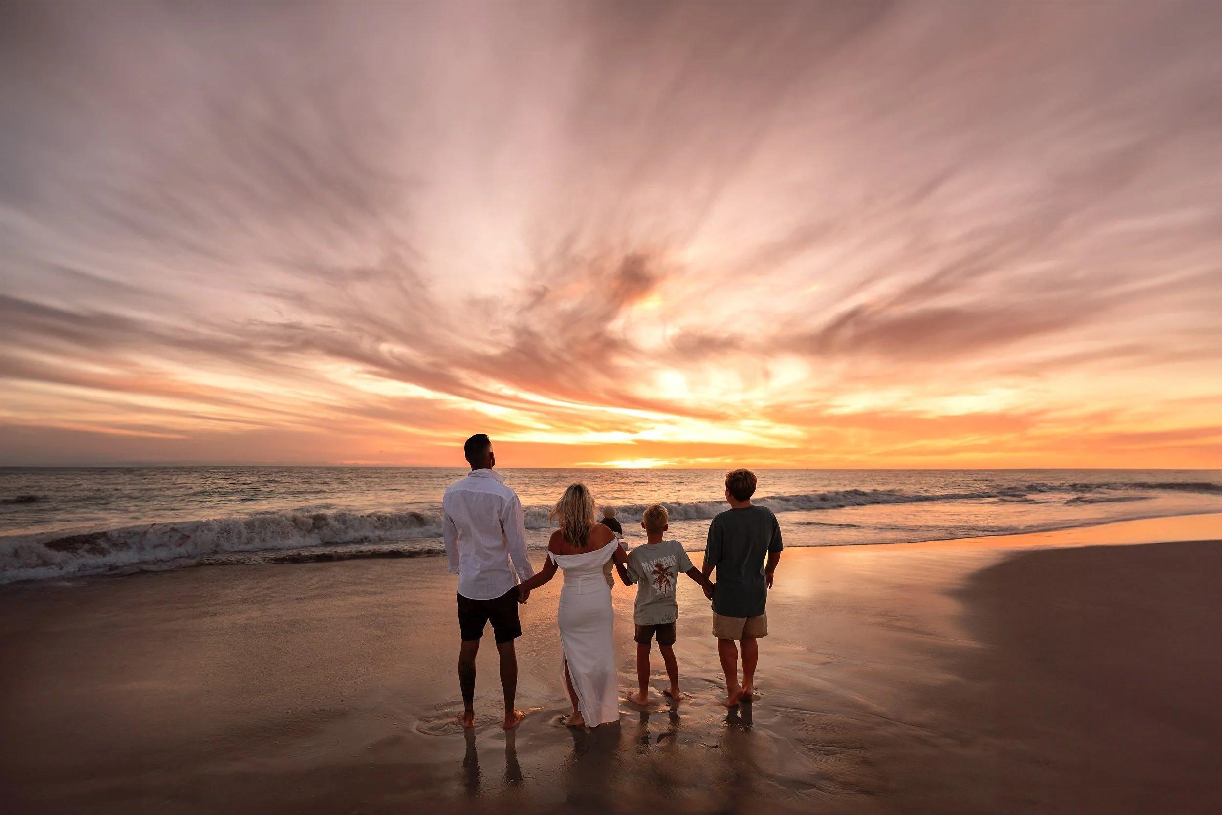 A group of five people, including two adults and three children, holding hands and walking on the beach during sunset.