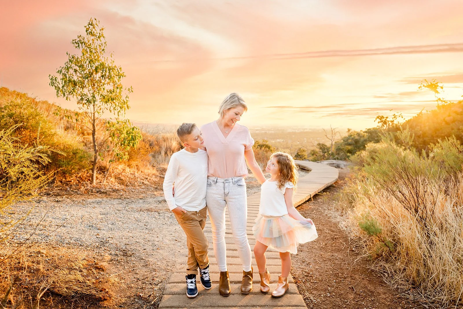 A woman and two children walking on a wooden trail during sunset, surrounded by dry grasses and trees.