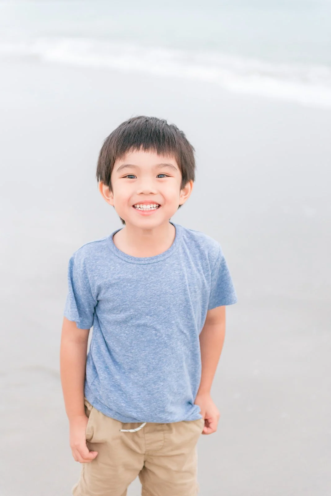 A young boy smiling at the camera on a beach with wet sand and gentle waves in the background.