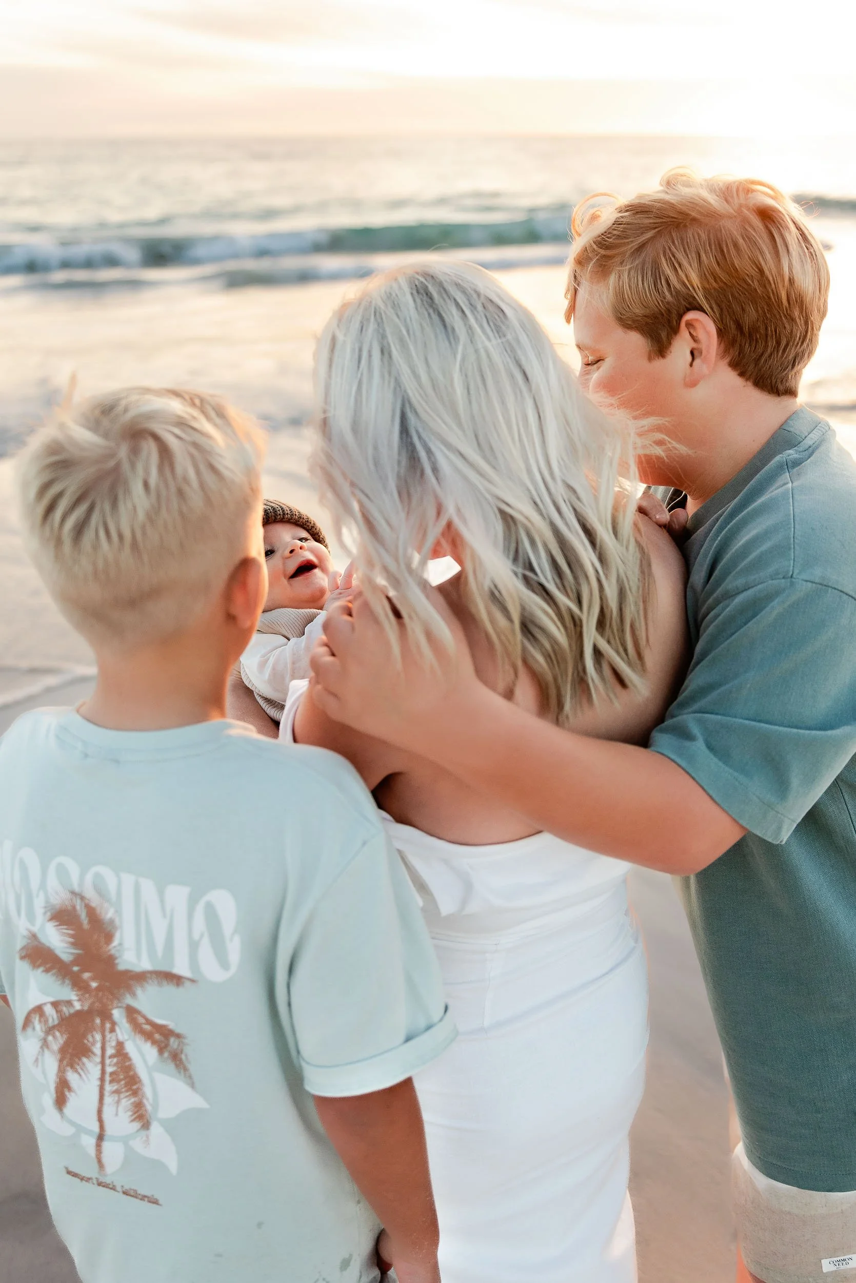 A family of four, including two young children, hugging and laughing on a beach at sunset.