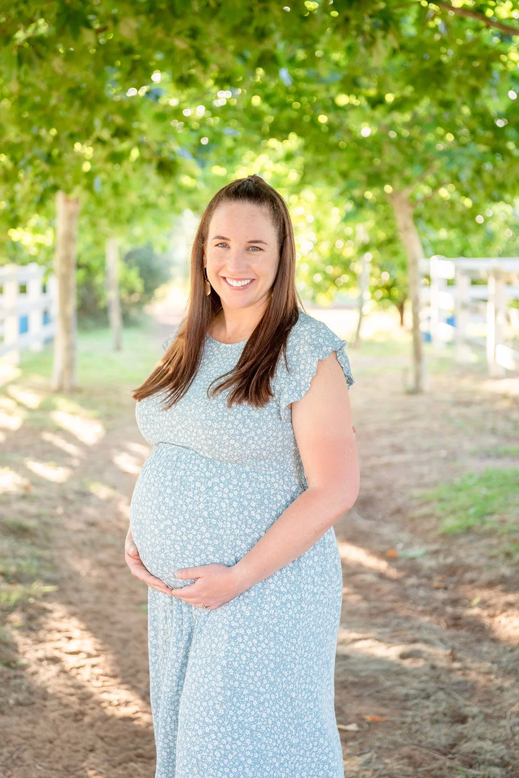 A pregnant woman with long brown hair, wearing a light blue dress with white floral pattern, standing outdoors on a dirt path surrounded by green trees and white fences, smiling.