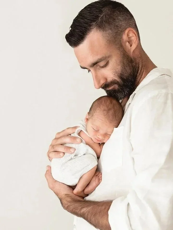 A man holding a sleeping newborn baby close to his chest, looking down at the baby with a gentle expression.