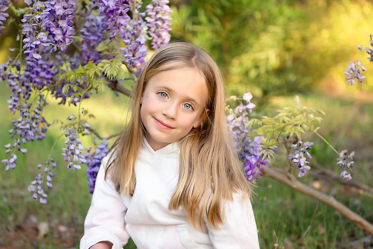 A young girl with long blonde hair and blue eyes smiling, sitting outdoors near purple wisteria flowers and green foliage.