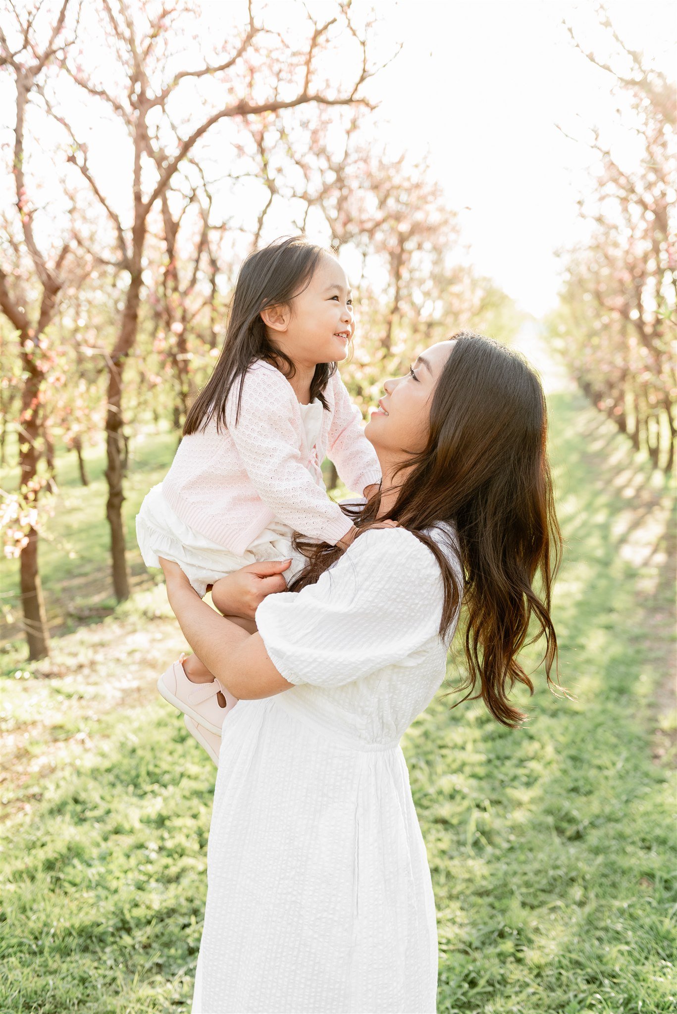 A woman is holding a young girl in a pink sweater and white skirt in a park or orchard with flowering trees. They are smiling and looking at each other as the sun shines brightly in the background.
