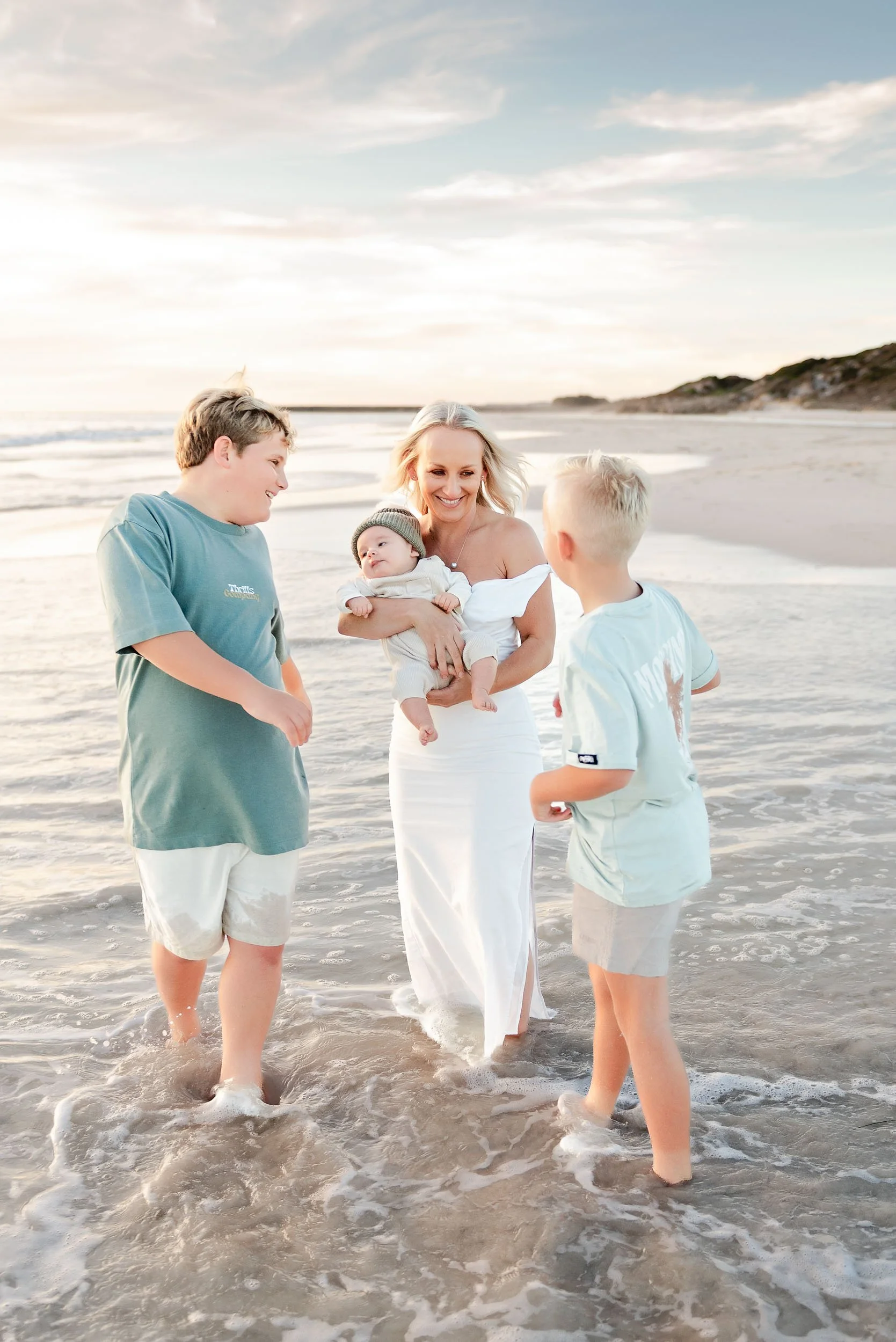 A woman holding a baby and playing with two young boys on the beach during sunset.