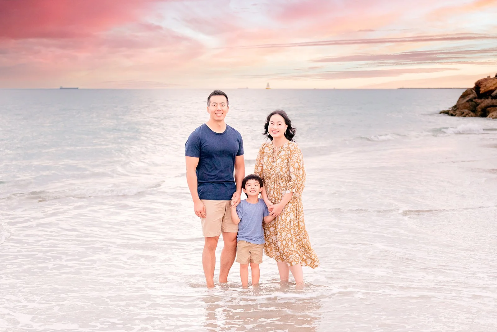 A family of three standing in shallow ocean water at sunset, smiling, with pink and orange sky and a rocky shoreline on the right.