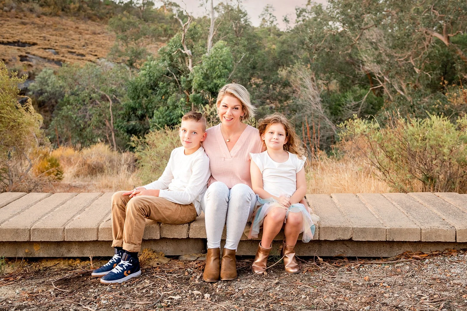 A woman with short blonde hair and two children sitting on a brick bench outdoors with trees and dry grass in the background.