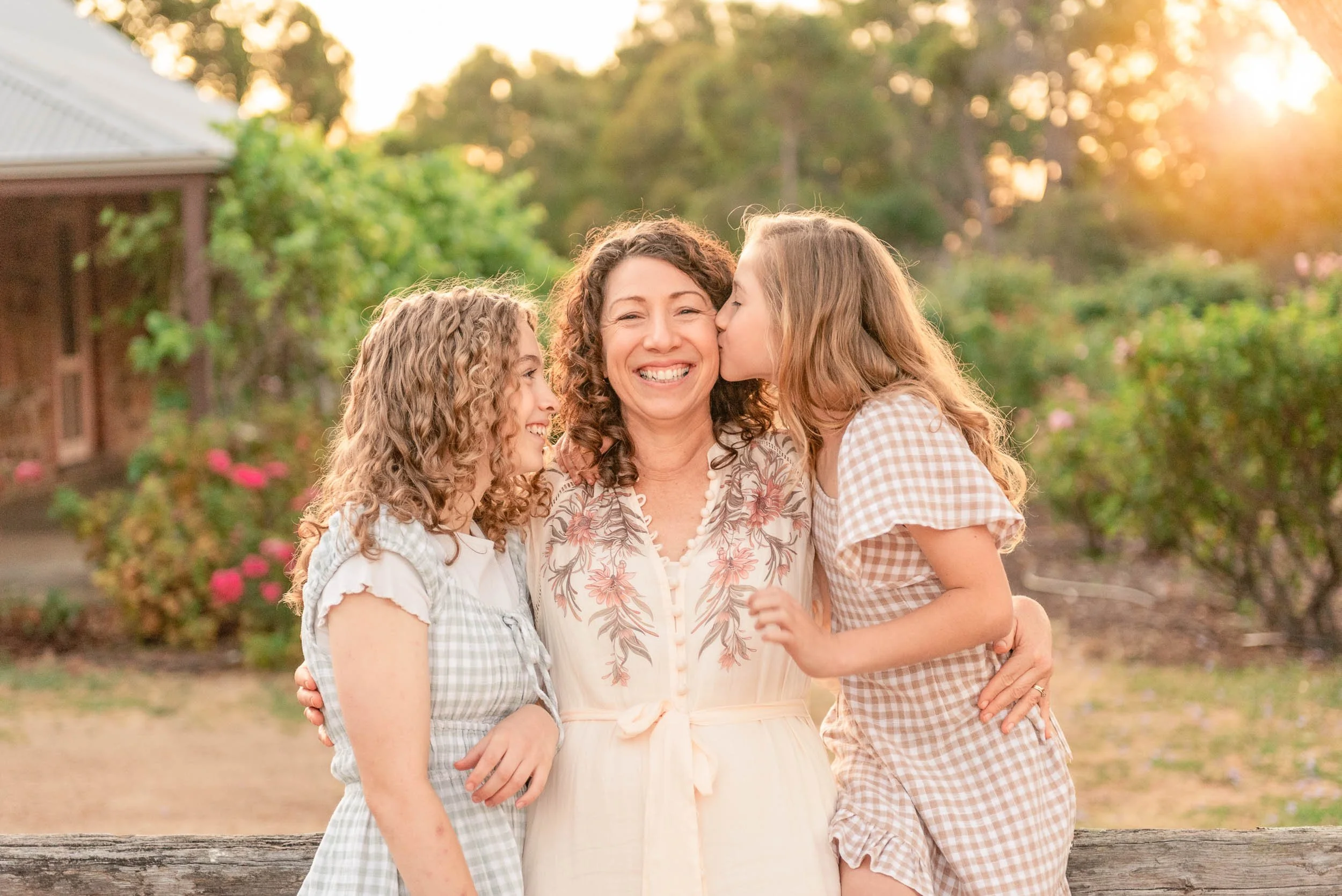 A woman with two young girls kissing her on the cheeks outdoors during sunset. All three are smiling and hugging each other, with trees and flowers in the background.