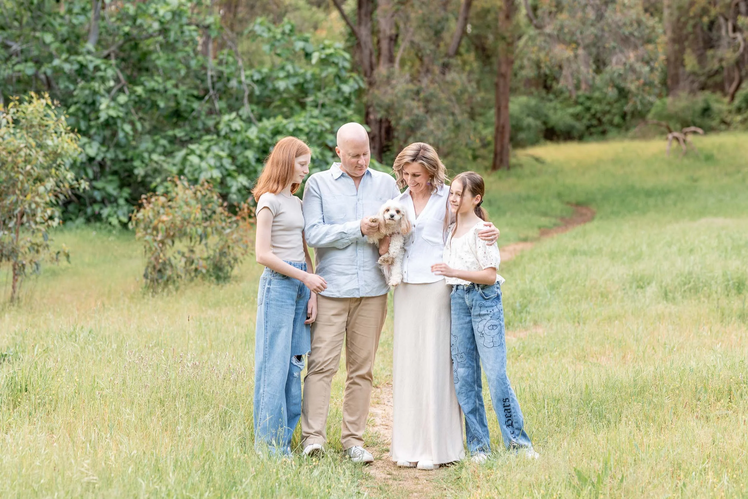 A family of five, including two young girls, an adult man and woman, and an elderly woman, standing on a grassy trail in a forest, holding a small white dog, with trees and greenery in the background.