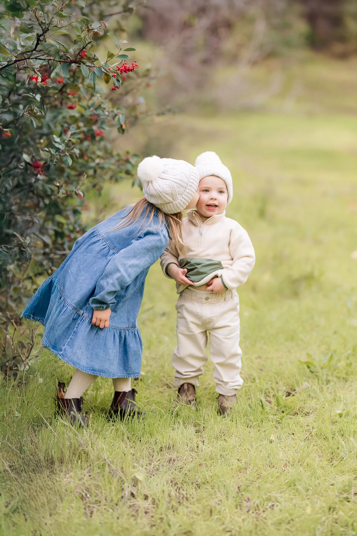Two young children, a girl and a boy, standing outdoors near a bush with red berries. The girl is whispering into the boy's ear. They are dressed warmly with knit hats, and the background is a grassy area with trees.