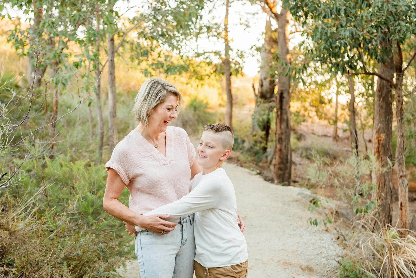 A woman and a boy hugging and smiling in a wooded park during sunset.