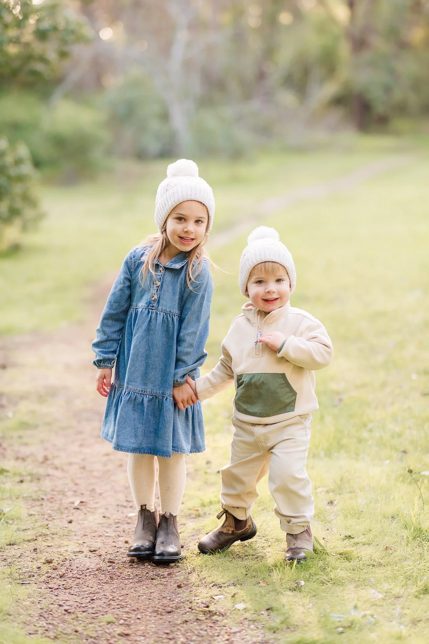 Two young children, a girl and a boy, holding hands and walking outdoors on a dirt path. They are dressed in warm clothing with white knit hats, and the background features trees and greenery.