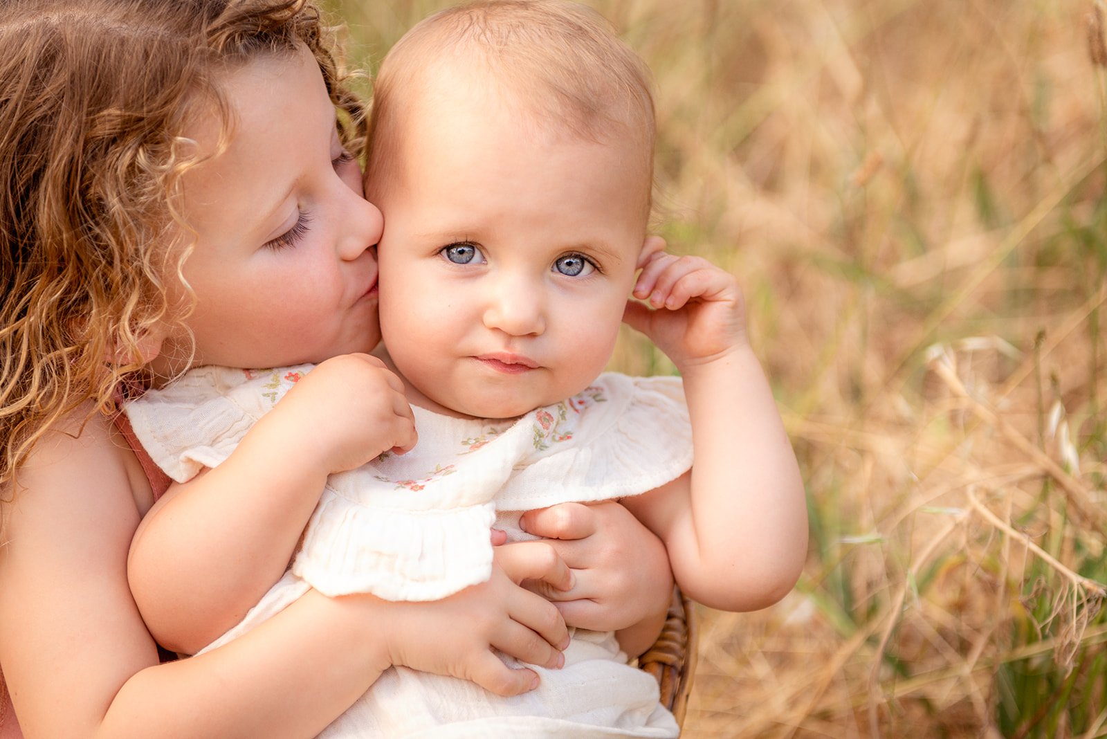 A young girl with curly red hair kisses a baby with blue eyes on the cheek. The baby has light hair and is wearing a white dress, while they are outdoors in a field with tall grass.